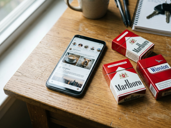 گوشی هوشمند کنار بسته‌های Marlboro، Camel و Winston برای نمایش تأثیر شبکه‌های اجتماعی بر ترند محصولات